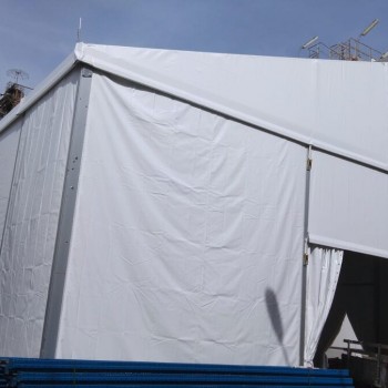 white tent structure at industrial storage tank site, truck visible, blue barrier pipes stacked, industrial towers in background