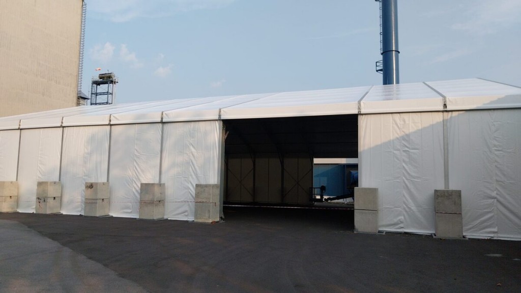 exterior white A-frame warehouse-style tent, open entrance, concrete block weights, industrial tower structure in background, daytime, empty