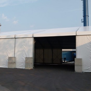 exterior white A-frame warehouse-style tent, open entrance, concrete block weights, industrial tower structure in background, daytime, empty