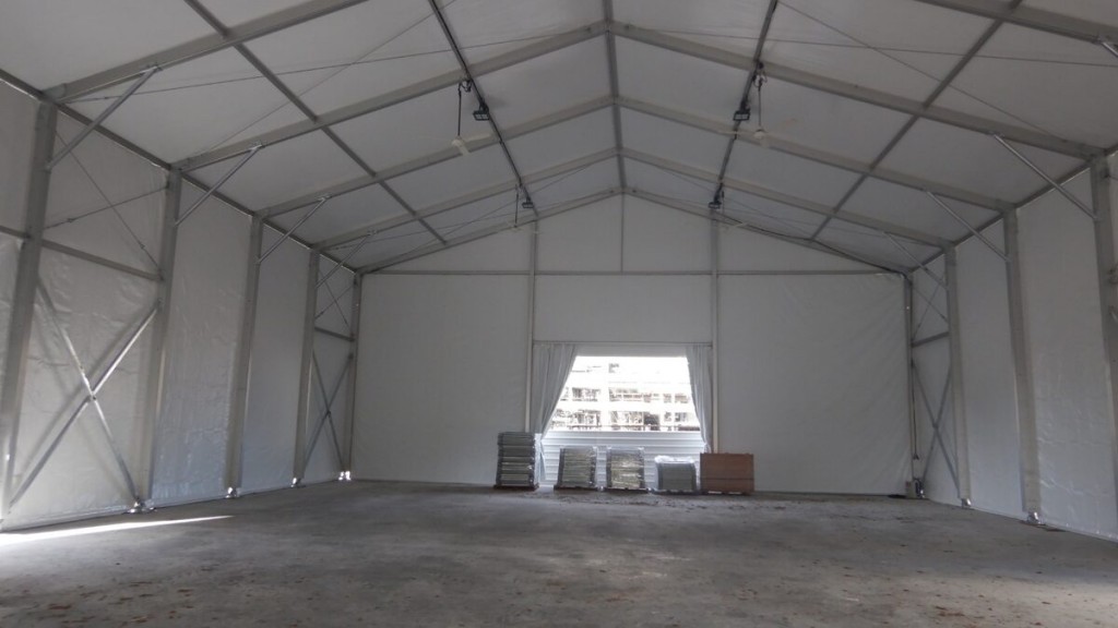 interior of large empty white aluminium tent, exposed truss ceiling, building visible through open end, concrete floor with construction materials