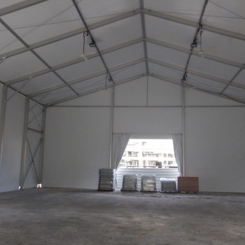 interior of large empty white aluminium tent, exposed truss ceiling, building visible through open end, concrete floor with construction materials