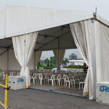 white A-frame tent with draped curtains, open sides showing white plastic tables and chairs inside, concrete ballast blocks, industrial coastal area in background