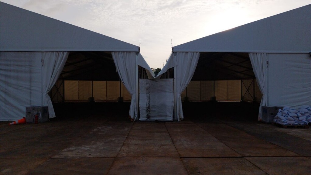symmetric view of two connected white tent structures meeting at peak, dusk sky, draped curtain entrances, outdoor industrial site with sandbags