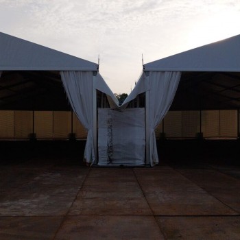 symmetric view of two connected white tent structures meeting at peak, dusk sky, draped curtain entrances, outdoor industrial site with sandbags