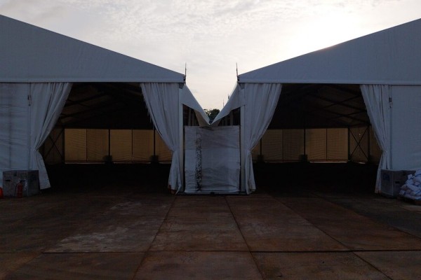 symmetric view of two connected white tent structures meeting at peak, dusk sky, draped curtain entrances, outdoor industrial site with sandbags