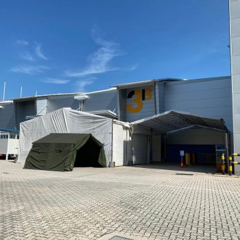 small dark green canvas dome tent set up against large industrial warehouse building exterior, paved yard, no people
