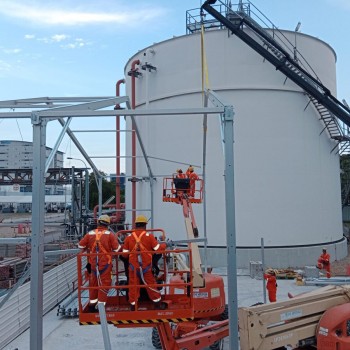 workers on cherry pickers installing structure near large white industrial storage tank, construction crew in orange coveralls