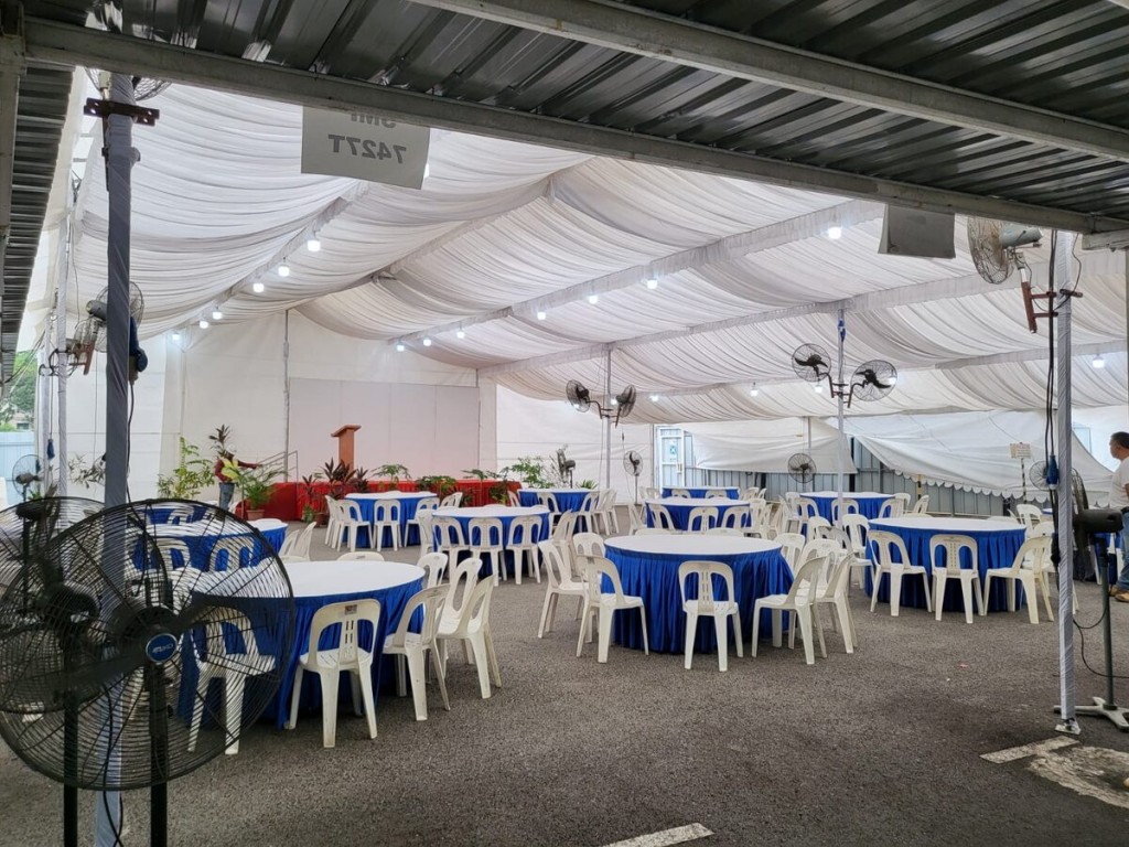 indoor tent event with blue-skirted round tables and white plastic chairs, draped ceiling, standing fans, empty venue before guests arrive