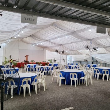 indoor tent event with blue-skirted round tables and white plastic chairs, draped ceiling, standing fans, empty venue before guests arrive