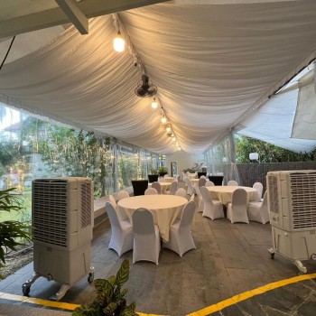 white draped tent with hanging string lights, round tables with white chair covers, two portable evaporative coolers, evening courtyard setting