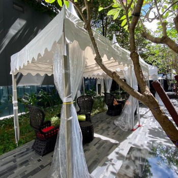 small white scalloped-edge canopy tent among trees in garden setting, wicker chairs with cushions underneath, boutique feel