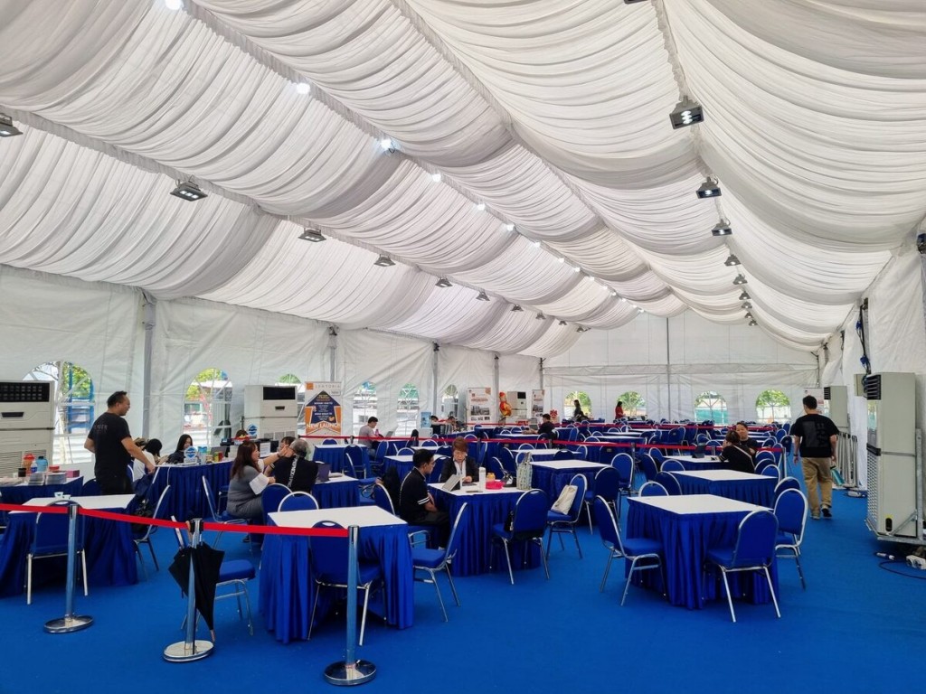 indoor tent event, blue-skirted round tables with white chairs, red rope queue divider, guests seated, draped white ceiling with pendant lights