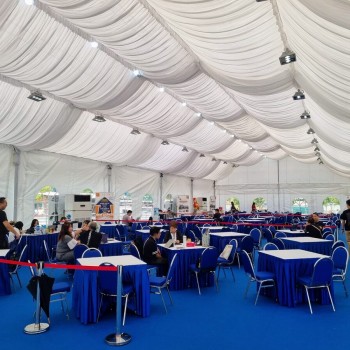 indoor tent event, blue-skirted round tables with white chairs, red rope queue divider, guests seated, draped white ceiling with pendant lights