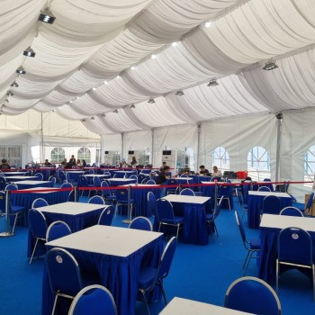 indoor tent event, blue-skirted square tables with white chairs, red rope queue barrier, draped ceiling, guests seated in background