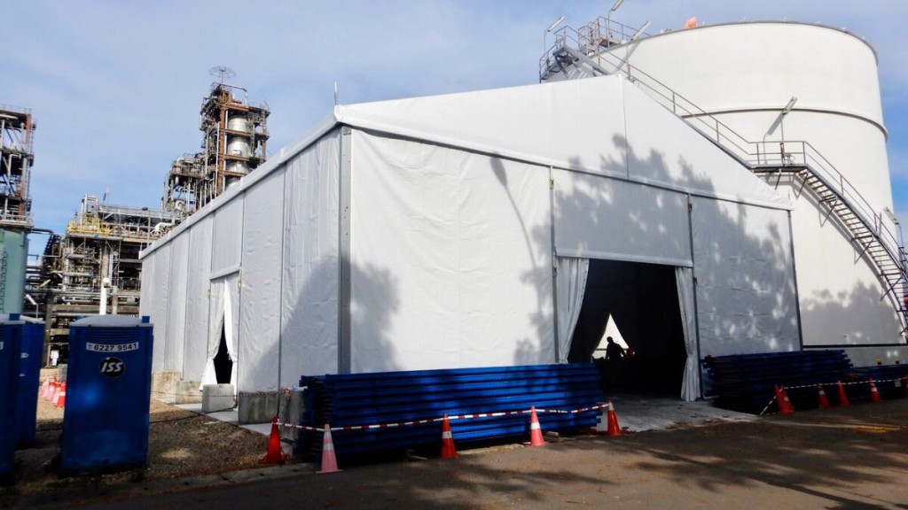 white tent set up alongside industrial storage tank silo, staircase visible, blue barrier pipes stacked, industrial site