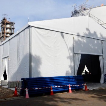 white tent set up alongside industrial storage tank silo, staircase visible, blue barrier pipes stacked, industrial site