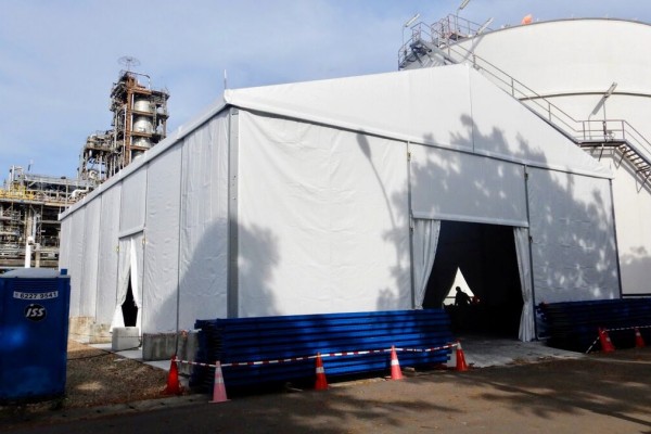 white tent set up alongside industrial storage tank silo, staircase visible, blue barrier pipes stacked, industrial site