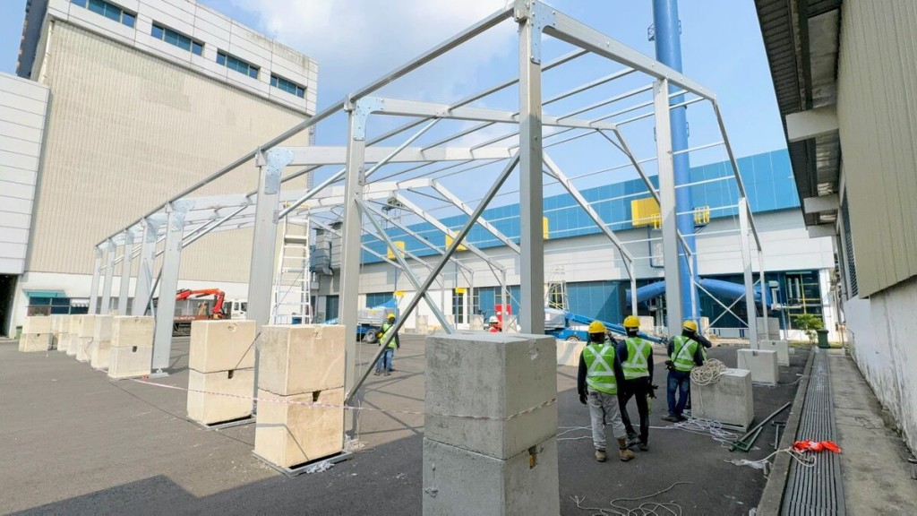 aluminium tent frame structure being erected outdoors, workers in high-vis vests and hard hats, industrial building backdrop, daytime