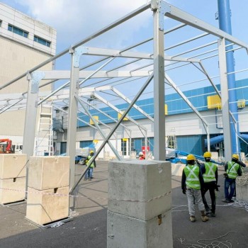 aluminium tent frame structure being erected outdoors, workers in high-vis vests and hard hats, industrial building backdrop, daytime