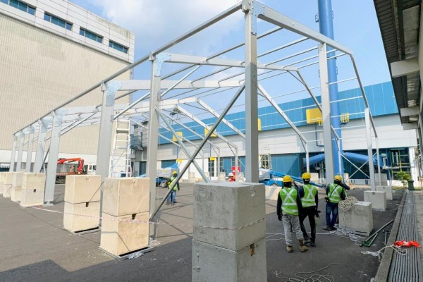 aluminium tent frame structure being erected outdoors, workers in high-vis vests and hard hats, industrial building backdrop, daytime