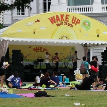 yellow branded promotional tent stage with sponsor signage, live band performing outdoors, people sitting on yoga mats on grass