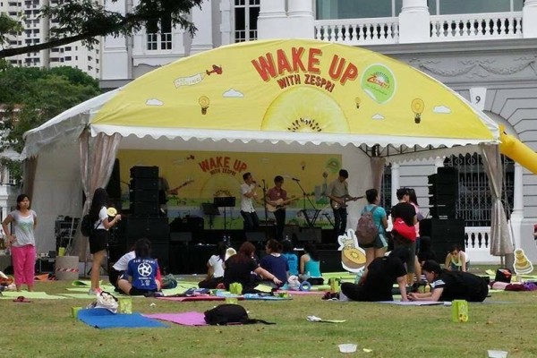yellow branded promotional tent stage with sponsor signage, live band performing outdoors, people sitting on yoga mats on grass