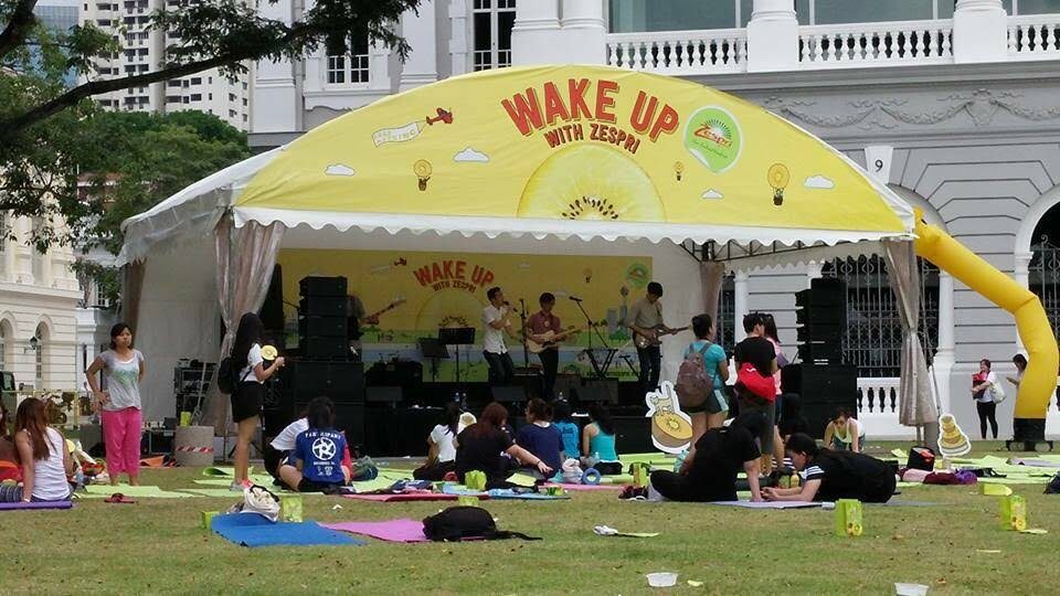 yellow branded promotional tent stage with sponsor signage, live band performing outdoors, people sitting on yoga mats on grass