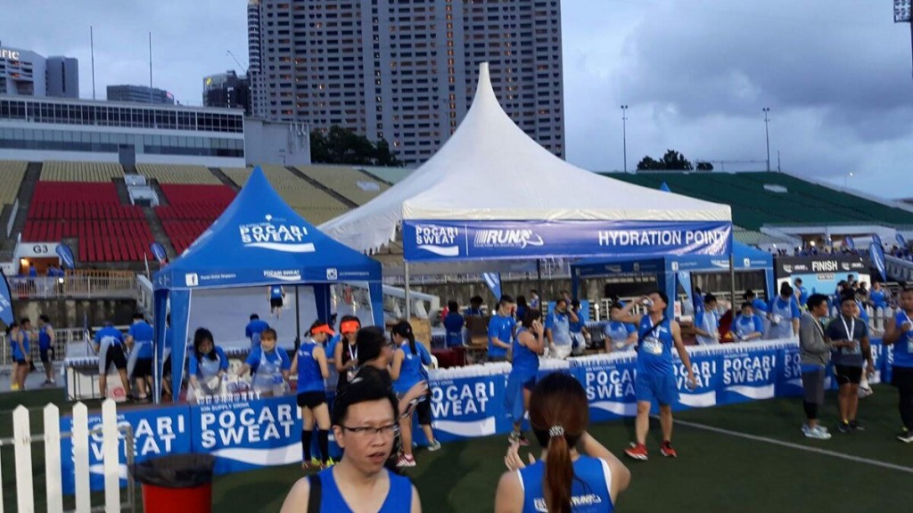 blue and white branded canopy tents at outdoor sports event hydration point, runners in blue shirts, stadium bleachers in background