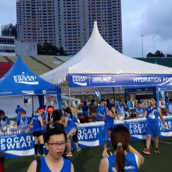 blue and white branded canopy tents at outdoor sports event hydration point, runners in blue shirts, stadium bleachers in background