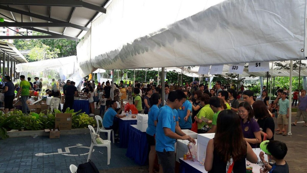 outdoor community bazaar under white tent canopy, crowd of people browsing $20 pricing sale tables, HDB void deck setting, daytime
