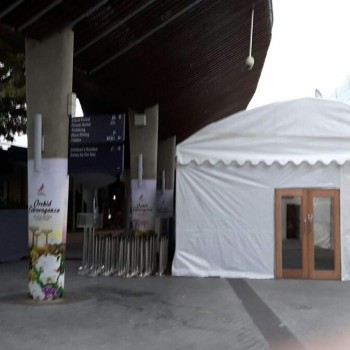 white A-frame tent with wooden double door entrance, outdoor plaza near modern building, orchid exhibition banners nearby