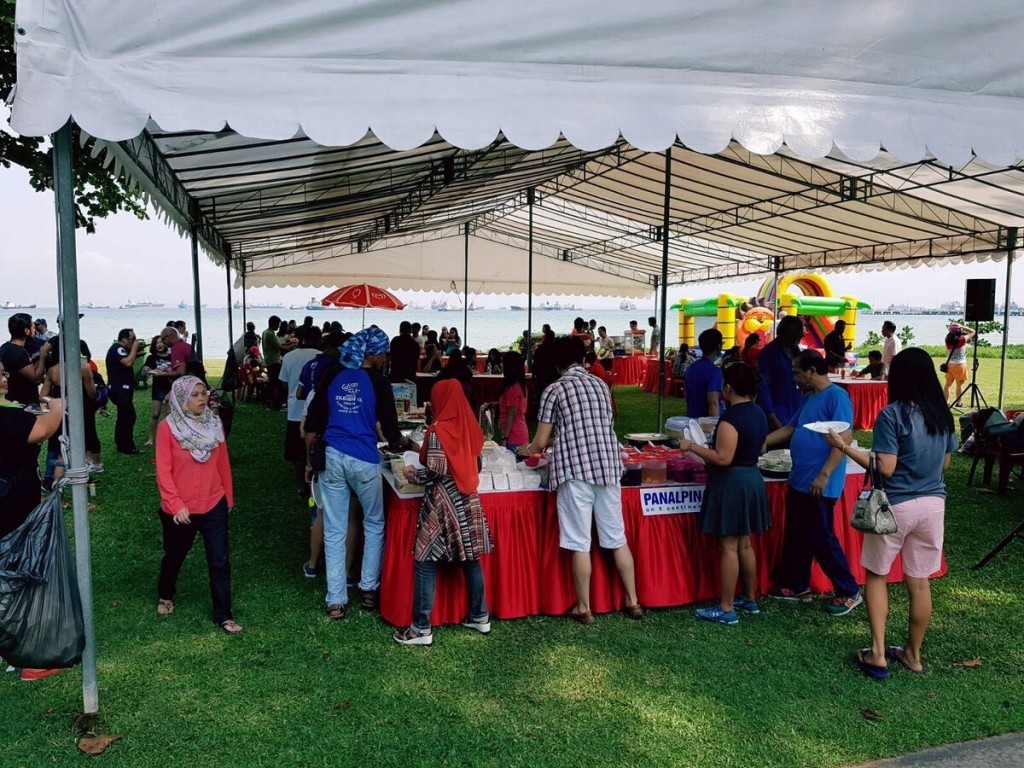 white peaked tent at outdoor waterfront community event, people gathered around red-skirted booth table, colourful inflatable in background