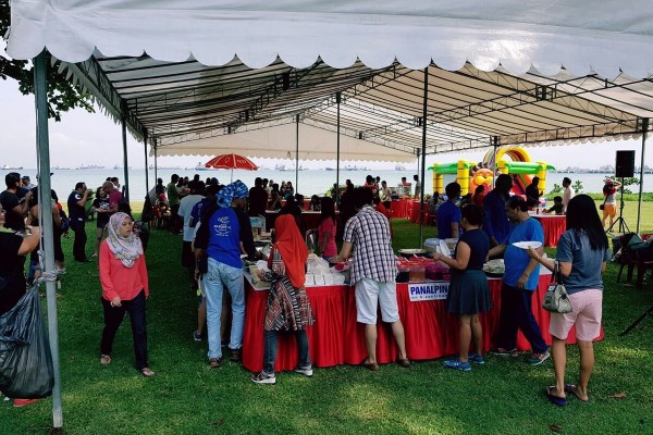 white peaked tent at outdoor waterfront community event, people gathered around red-skirted booth table, colourful inflatable in background