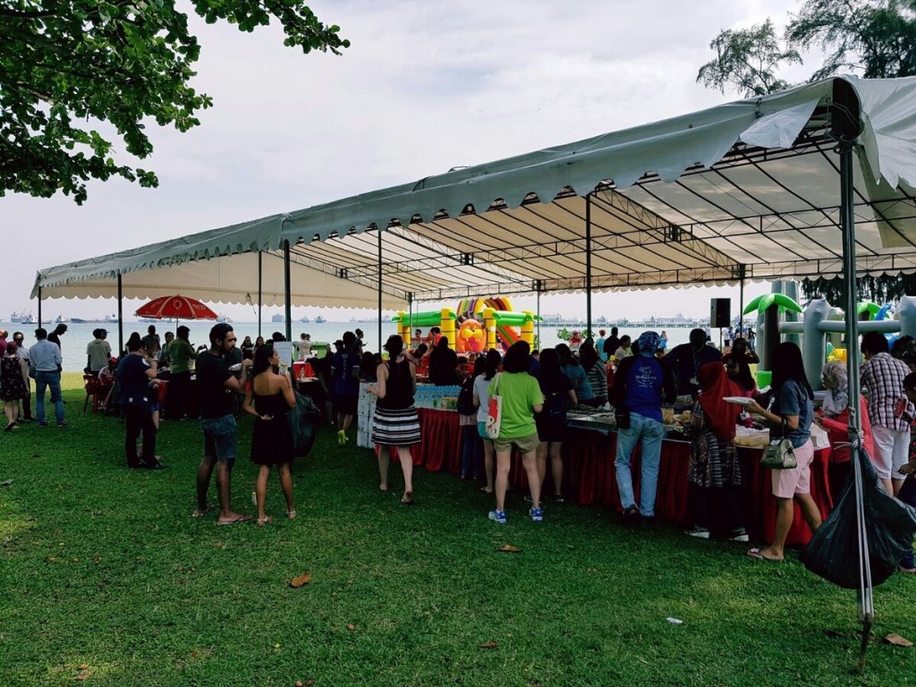 white outdoor waterfront event tent with crowd gathered for buffet, grass field near sea, colourful inflatable in background