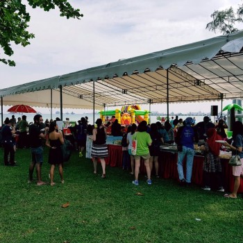 white outdoor waterfront event tent with crowd gathered for buffet, grass field near sea, colourful inflatable in background