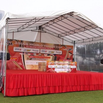 small pyramid tent with clear PVC sides, red-skirted promotional booth inside with sports sponsor banners, outdoor park setting near city skyline