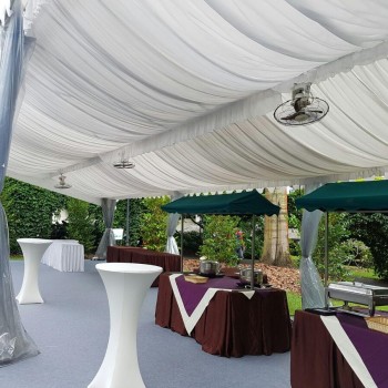 white draped tent with standing fan, cocktail tables with dark tablecloths, buffet table setup, outdoor garden setting