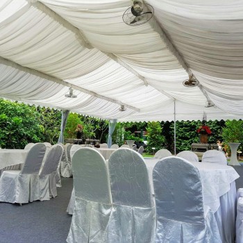 white draped tent ceiling with standing fan, rows of satin white chair covers, outdoor garden setting, empty before event