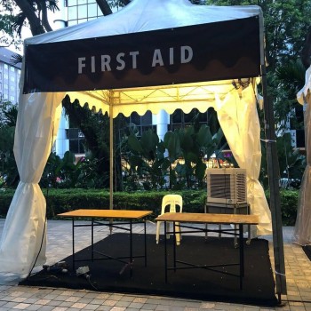white pagoda tent with first aid branded banner, yellow table and chair, portable aircon, garden setting near office building