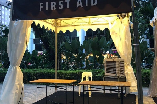 white pagoda tent with first aid branded banner, yellow table and chair, portable aircon, garden setting near office building