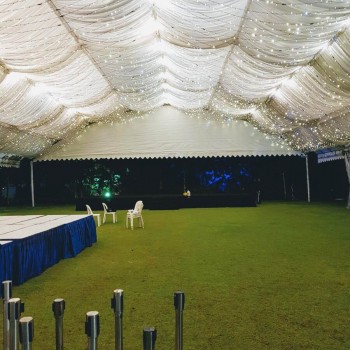 white draped tent ceiling with string fairy lights, empty grass field, blue-clothed table and stacked chairs, tiki torches lining ground, night setting