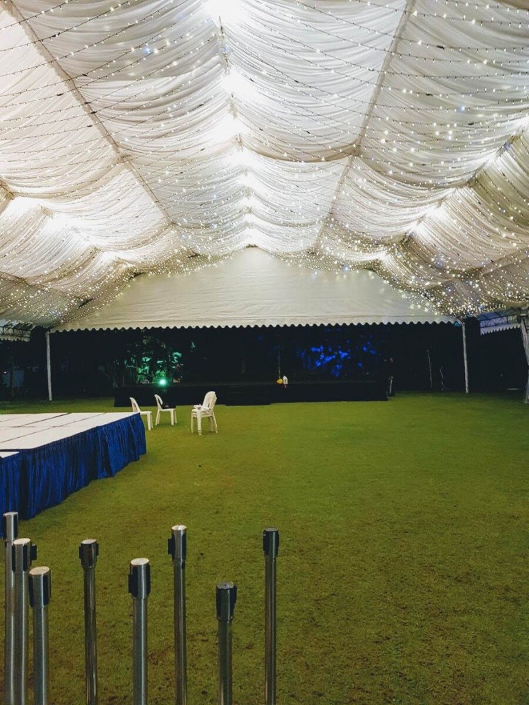 white draped tent ceiling with string fairy lights, empty grass field, blue-clothed table and stacked chairs, tiki torches lining ground, night setting