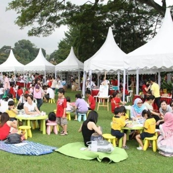 multiple white pagoda tents at outdoor community family event, people at yellow tables with children, grass field daytime