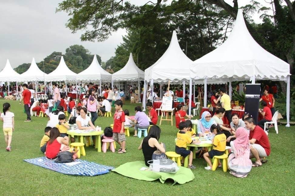 multiple white pagoda tents at outdoor community family event, people at yellow tables with children, grass field daytime