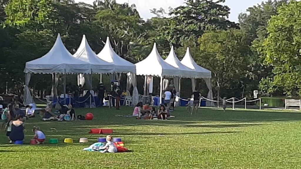 row of white pagoda tents on grass field in park, people relaxing with children, daytime picnic setting