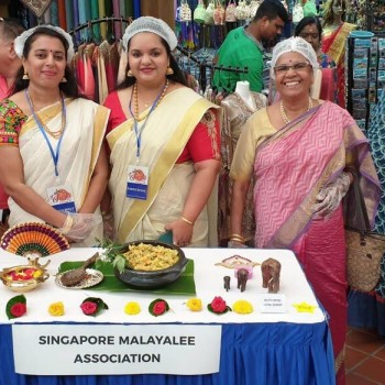 indoor community bazaar booth with association banner, women in traditional attire and headscarves at food and craft display table
