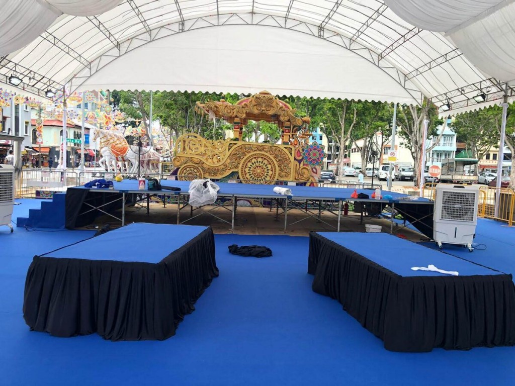 white draped tent stage setup with blue-clothed platforms, portable evaporative cooler, ornate decorative float prop, outdoor community event stage