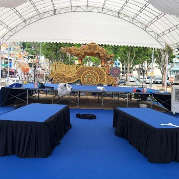 white draped tent stage setup with blue-clothed platforms, portable evaporative cooler, ornate decorative float prop, outdoor community event stage