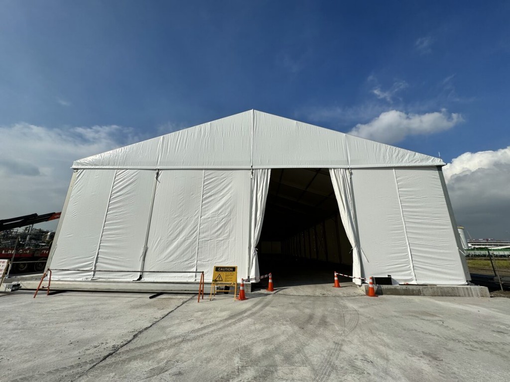 outdoor daytime white A-frame tent exterior, open entrance, traffic cones, car park area in background, empty, no people