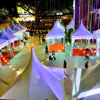 aerial view of purple-lit pagoda tents at night market bazaar, colourful string lights, people walking between tents